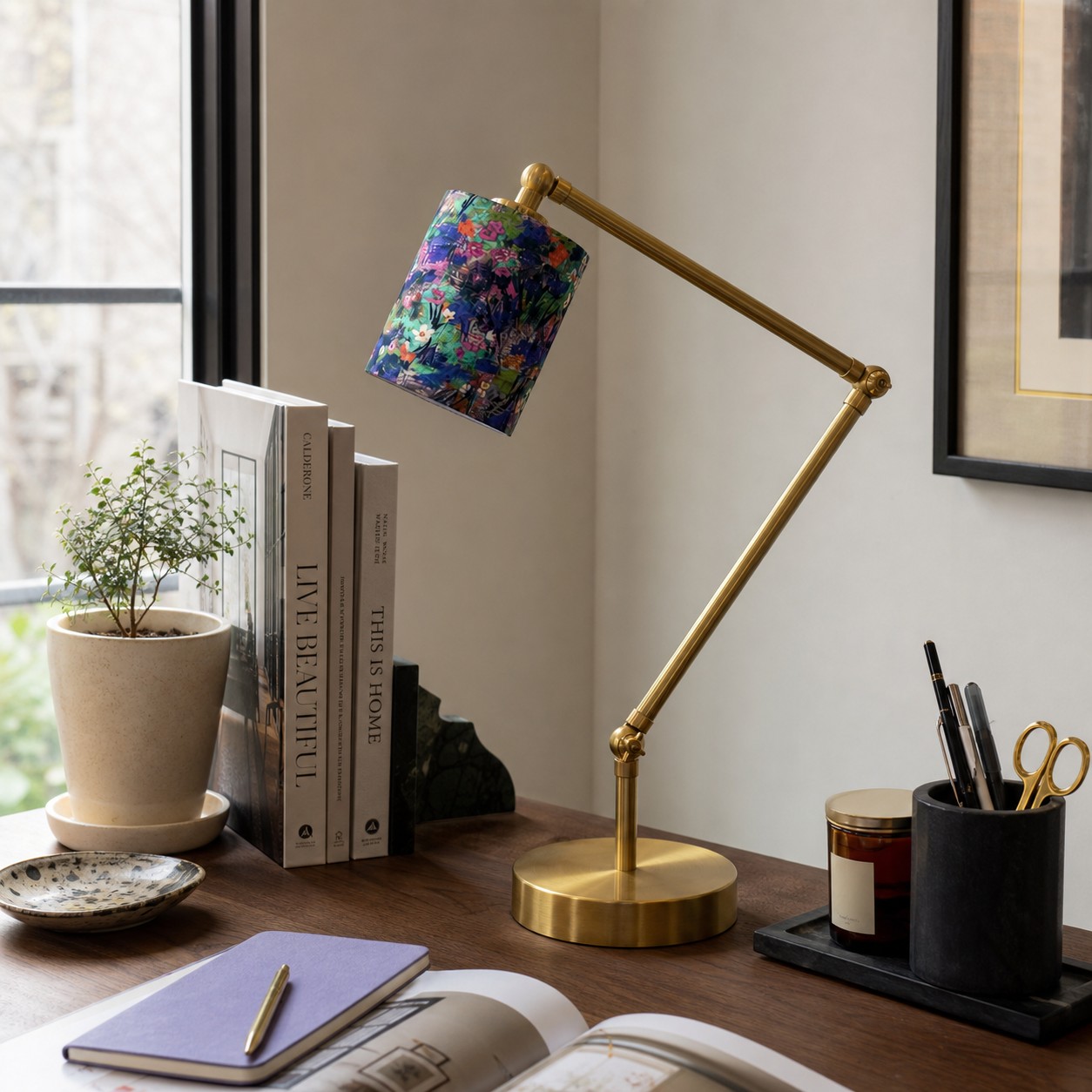 Eine Tischlampe mit floralem Schirm steht auf einem Schreibtisch neben Büchern und Dekoartikeln.